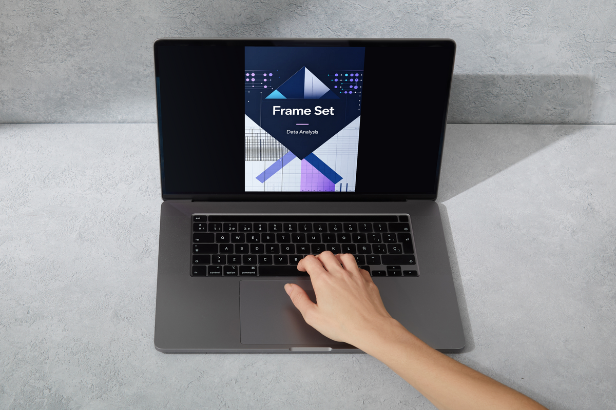 A gray laptop on a light gray textured surface, with a hand resting on the trackpad. The screen displays a book or course cover titled 'Frame Set' with geometric graphics.